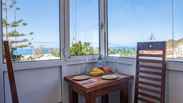 A balcony with a wooden table, two chairs, and a bottle of wine. The view includes a beach and buildings. - Inviting Arokaria Apartment in Panormo (Mylopotamos)