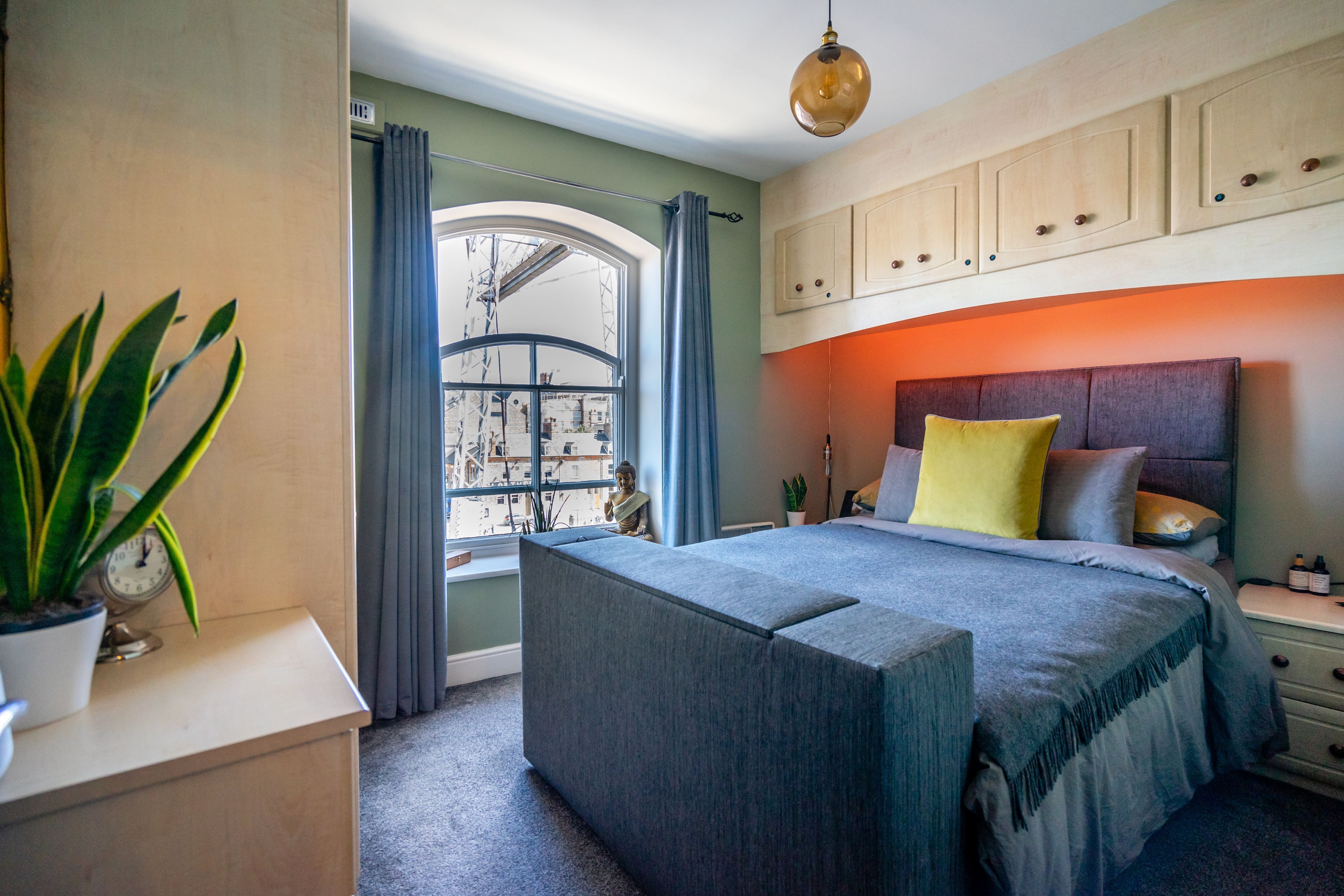 A bedroom with a bed, a bedside table, a nightstand, a window with curtains, and a plant.
