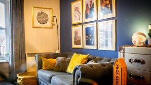 Una habitación con un sofá gris, cojines amarillos, una maleta antigua y cuadros en la pared.