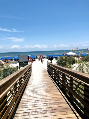 Une promenade en bois mène à une plage où se trouvent des parasols bleus et un panneau indiquant les nouvelles règles applicables à la plage.