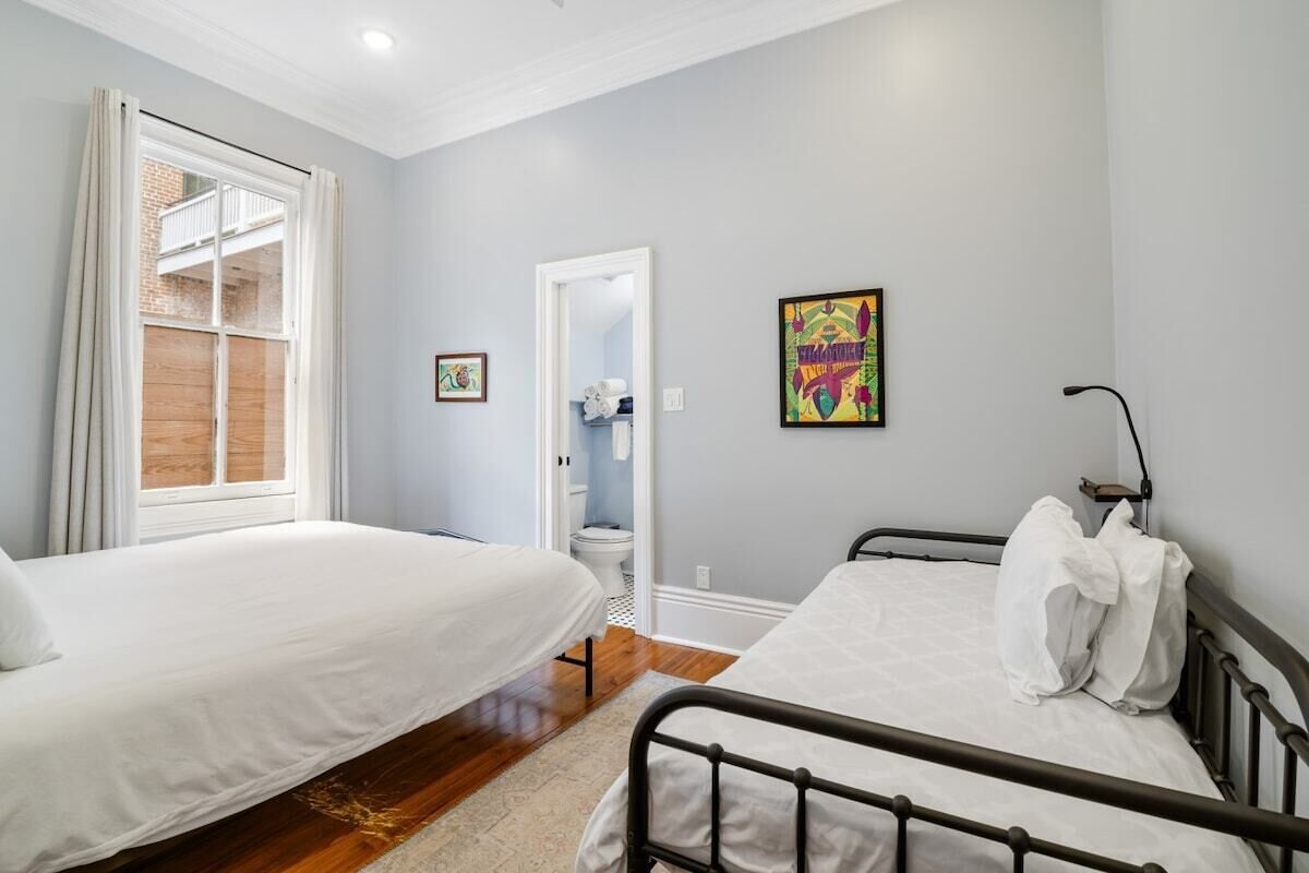 A bedroom with two beds, a window with curtains, a door leading to a bathroom, and a colorful painting on the wall.