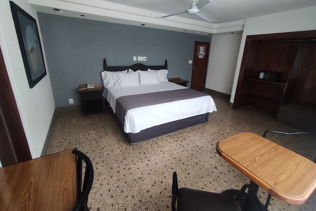 Una habitación de hotel con cama, escritorio, silla, ventilador de techo, mesita de noche y armario.