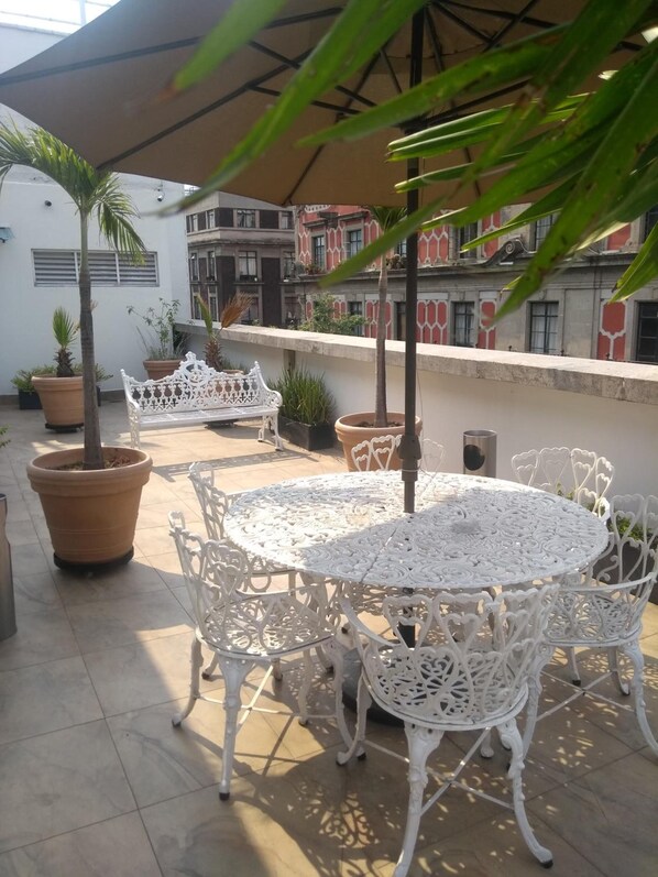 Un patio en la azotea con mesas y sillas de metal blanco ornamentado, rodeado de plantas en macetas y un paraguas grande.