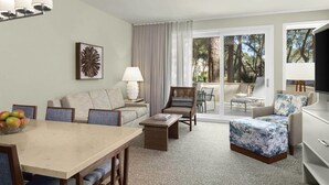 Un salon moderne comprenant un canapé, des fauteuils, une table basse et une table à manger. Une grande fenêtre offre une vue sur les arbres, et il y a un balcon.