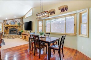 A dining area with a stone fireplace, a TV mounted on the wall, and large windows.