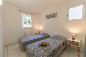 A small, well-lit bedroom with two beds, a bedside table with a lamp, and a window with a view of greenery.
