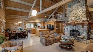 A log cabin interior with a stone fireplace, wooden furniture, and a kitchen area. - Eagle's Landing- Beach Front Log Home  - RARE FIND on Beautiful Woman Lake! (Longville)