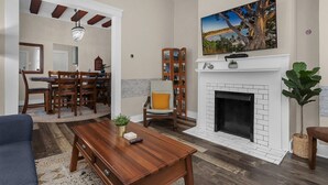 A living room with a fireplace, a television mounted above it, a dining area with a wooden table and chairs, and a large plant in the corner. - Modern❤️2BR near DT, 8 min walk to Franklin Park/Conservatory/Trolley District (Columbus)