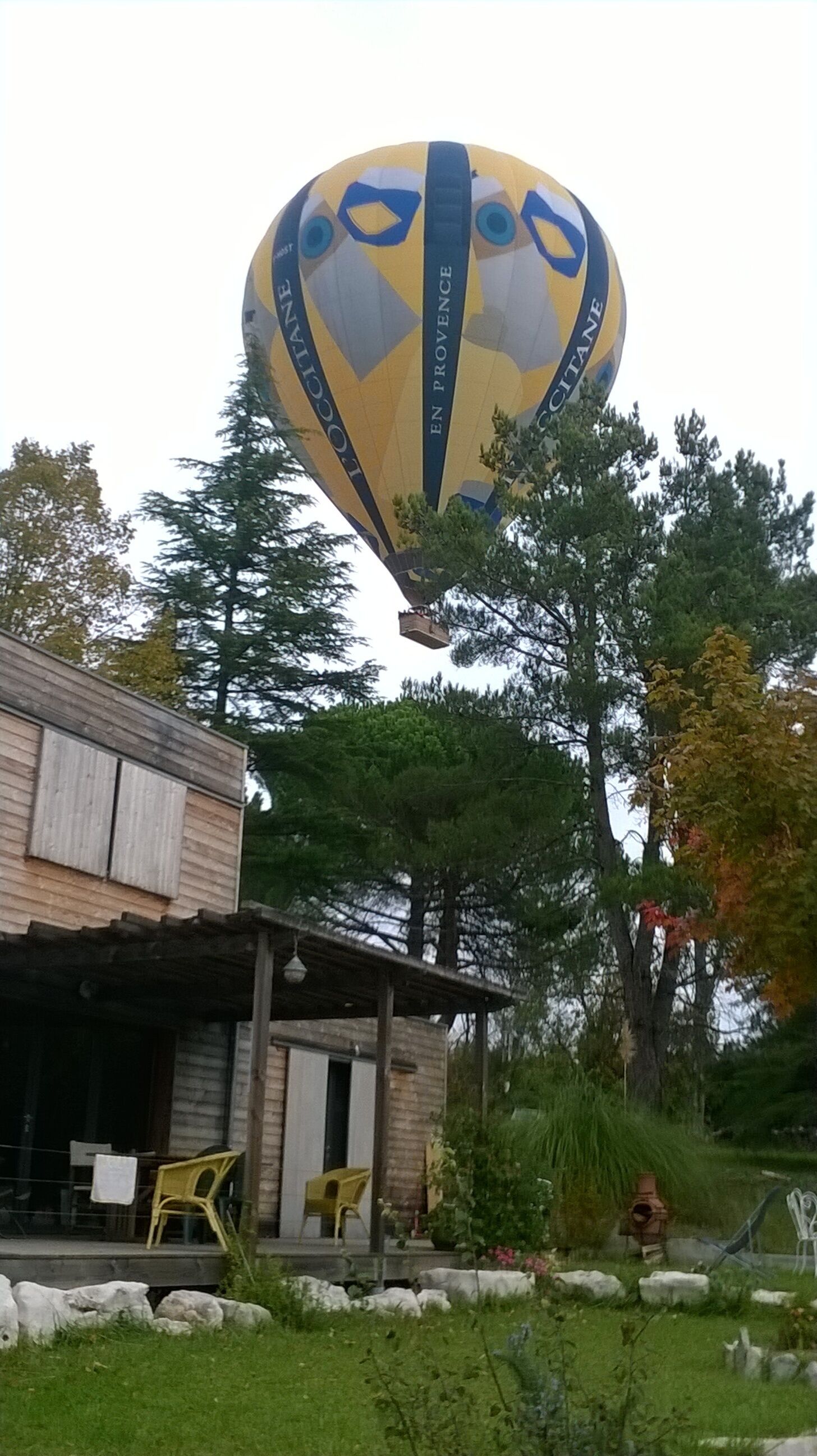 Un globo aerostático con la inscripción "EN PROVENCE" sobrevolando un edificio con porche y terraza.