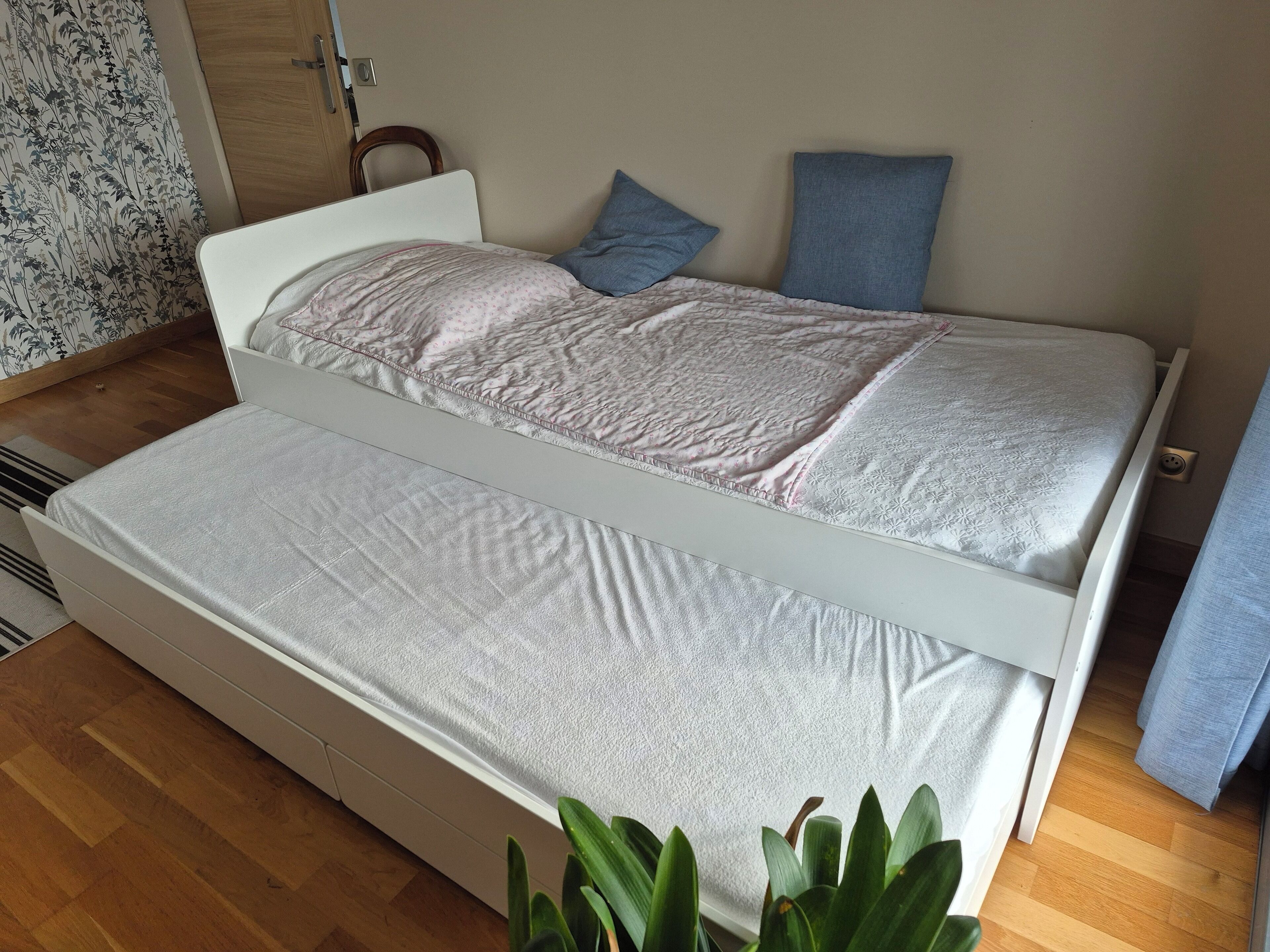 A single bed with a white frame and a grey mattress, two pillows, and a blue pillow on a wooden floor.