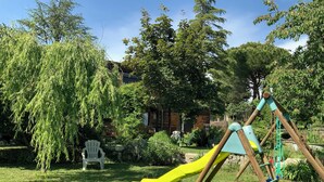 Un parc infantil de fusta amb tobogan groc en un jardí amb gespa, arbres i una casa de fusta al fons.