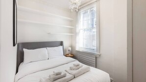En pænt redt seng med hvide sengetøj og håndklæder, en grå sengegavl, en sengetabletlampe og et vindue med rullegardiner.