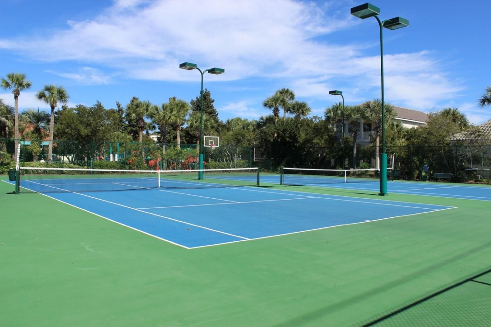 An outdoor tennis court with green and blue surfaces, surrounded by trees and a building in the background.