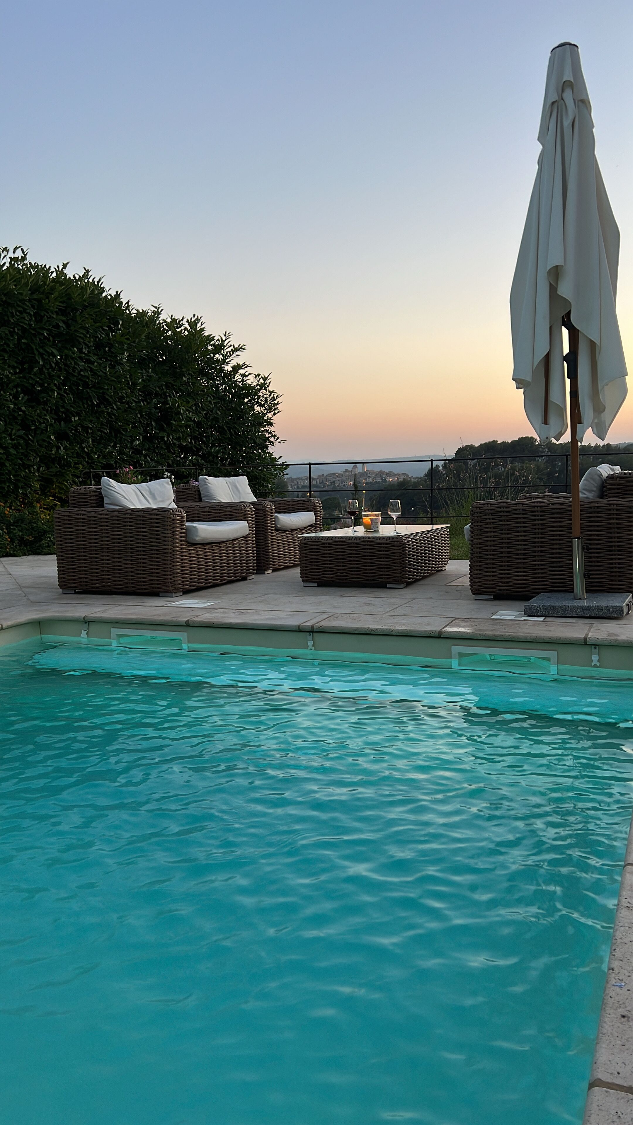 Un espace aménagé au bord de la piscine, avec des fauteuils de relaxation en osier et une table, un grand parasol, et une vue sur le lointain paysage urbain au coucher du soleil.