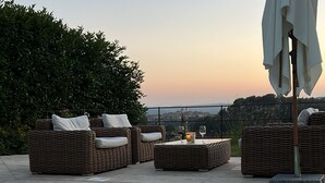 Zona junto a la piscina con sillones de mimbre, una mesa y una sombrilla grande, con vistas a un horizonte urbano al atardecer.