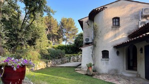 Un edificio rústico con un sendero de piedra que lleva a una puerta, rodeado de exuberante vegetación y una maceta grande con flores moradas.