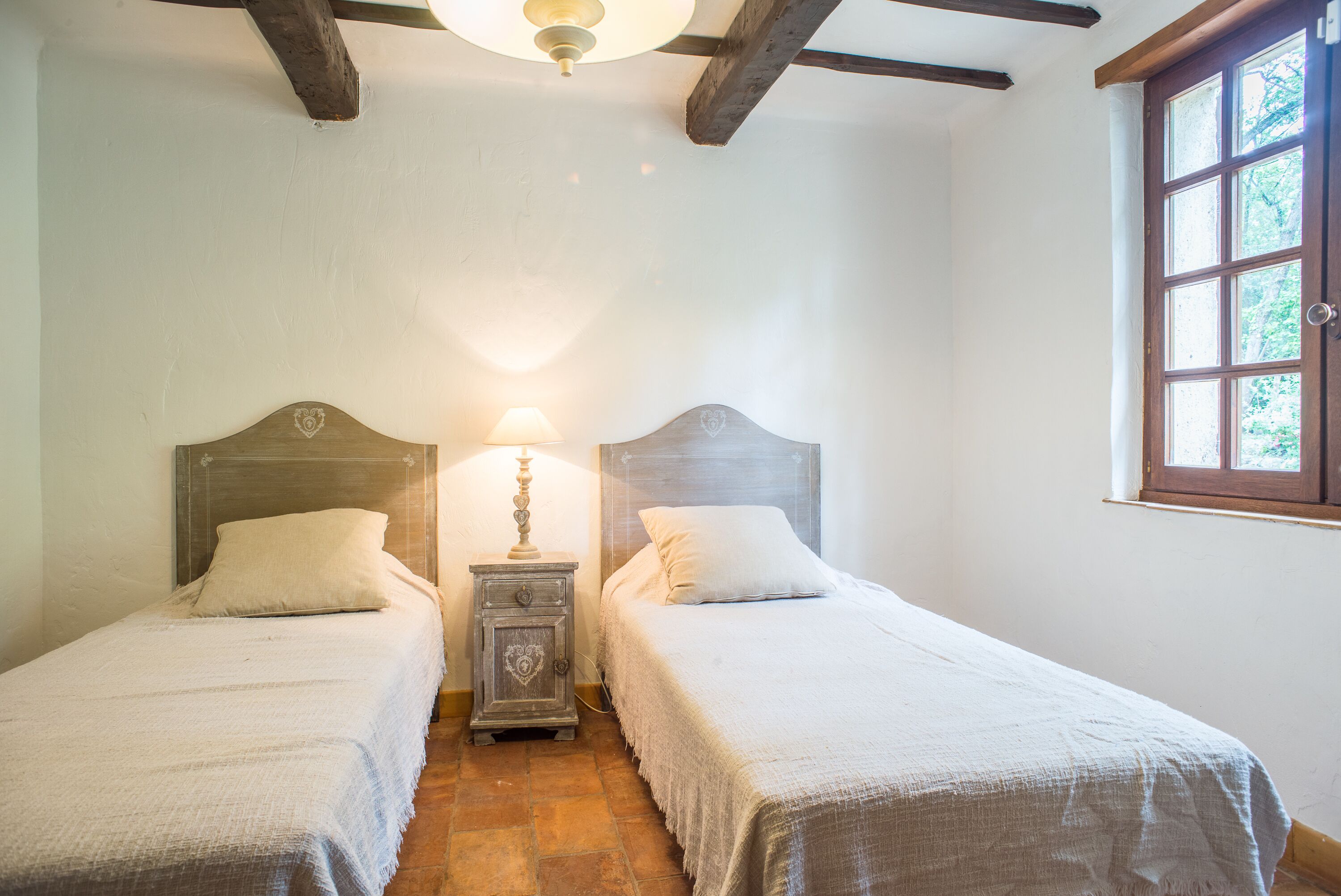 A bedroom with two single beds, a nightstand with a lamp, and a window with a view of trees.