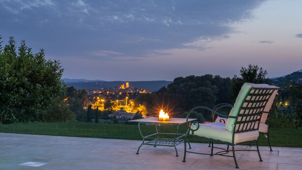 Un área de estar al aire libre con mesa y sillas, con vistas a un pueblo con edificios iluminados a lo lejos.