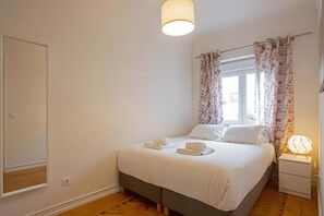 Ein Schlafzimmer mit Bett, Nachttisch, Lampe, einem Fenster mit Vorhängen und einem Spiegel an der Wand.