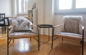 Zwei Korbsessel mit Felldecken, ein kleiner runder Tisch und ein Bücherregal in einem Raum mit Holzboden und großen Fenstern.