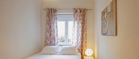 Un dormitorio con cama, cortinas, una ventana, un cuadro colgado en la pared y una lámpara de noche.