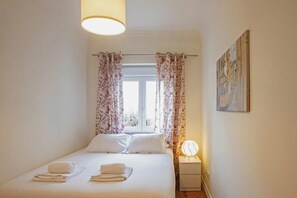 Een slaapkamer met een bed, gordijnen, een raam, een aan de muur hangend schilderij en een bedlamp.