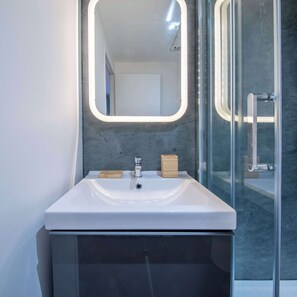 Ein Badezimmer mit einem weißen Waschbecken, einem Spiegel mit Beleuchtung, einer hölzernen Seifenschale und einer Glasdusche.