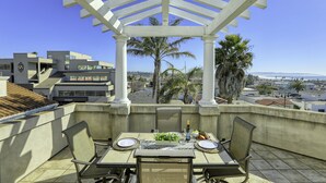 Une terrasse sur le toit, avec une table dressée pour deux personnes, entourée de chaises, et protégée par une pergola blanche.
