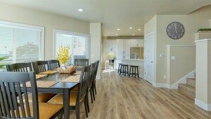 Une cuisine moderne avec des meubles blancs, un coin repas doté d’une table et de chaises en bois, et une grande horloge murale.