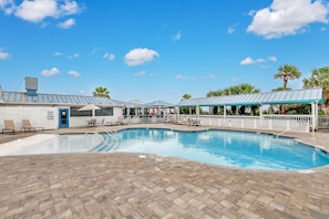 A swimming pool with lounge chairs and umbrellas, a covered seating area, and a building with a blue door.