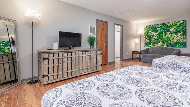 Quarto moderno com cômoda de madeira, TV de tela plana, sofá cinza e uma grande obra de arte com padrão de folhagem tropical.