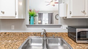 Cozinha com pia de aço inoxidável dupla, bancada de granito, armários brancos, um vaso azul-esverdeado com plantas, ventilador de teto e micro-ondas.