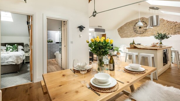 A modern kitchen with a wooden table set for dining, a stone wall, and a hanging light fixture. - Luxury Penthouse - Luxury Cottages (Cheltenham)