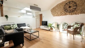 A modern living room with a black sofa, a coffee table, and a TV. There's a stone wall with a clock and a window with a view of the outside. - Luxury Penthouse - Luxury Cottages (Cheltenham)