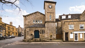 A historic stone building with a clock tower, a central door, and a small staircase leading to the entrance. - Luxury Penthouse - Luxury Cottages (Cheltenham)