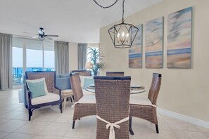 Un vaste espace de vie comprenant un coin repas avec une table entourée de chaises en osier, un ventilateur de plafond et une grande fenêtre donnant sur l’extérieur.