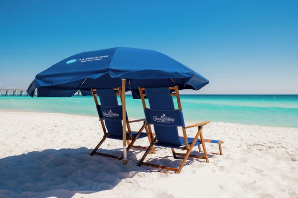 Dos sillas de playa con una sombrilla azul con la leyenda 'Beach Colony' en una playa arenosa.