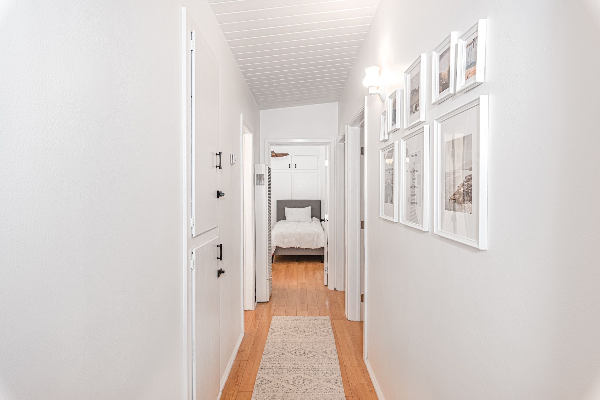 Un couloir étroit aux murs blancs et au plancher en bois, qui mène à une chambre à coucher équipée d’un lit et d’une armoire.