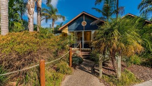 A tropical resort with a wooden deck leading to a glass entrance, surrounded by lush greenery and palm trees. - In Da Block - Beachy Haven (Inverloch)