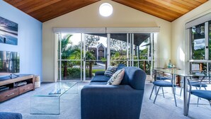 A modern living room with a large glass door leading to a balcony, a television, and a dining area with chairs. - In Da Block - Beachy Haven (Inverloch)