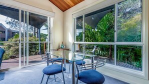 A conservatory with a glass table and chairs, surrounded by large windows offering a view of lush greenery. - In Da Block - Beachy Haven (Inverloch)