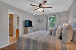 Un dormitorio con cama, ventilador de techo, televisor y ventana con cortinas.
