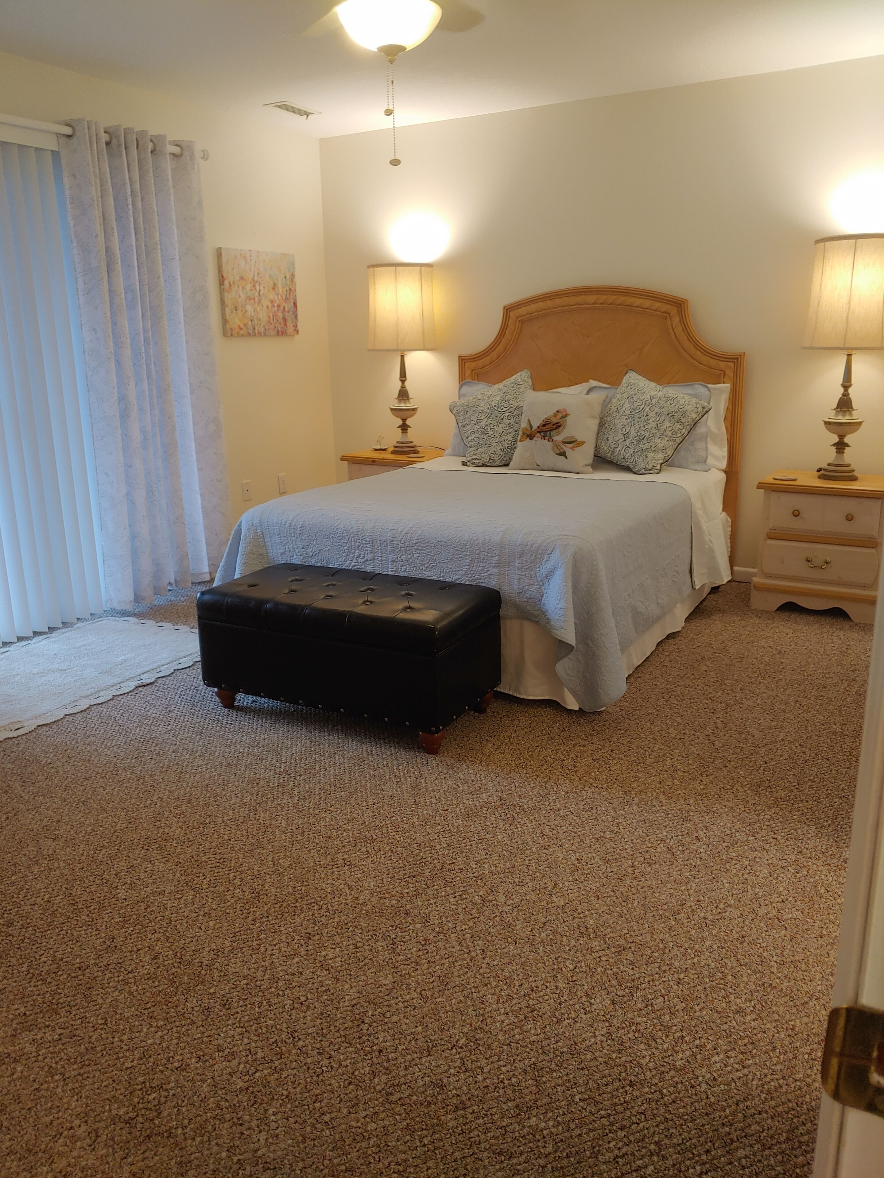Quarto com uma cama, duas mesas de cabeceira com luminárias, uma cabeceira de madeira, um banquinho preto e uma janela grande com cortinas.