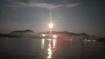 Vista noturna de uma massa de água com montanhas ao longe e uma fonte de luz brilhante refletindo na água.