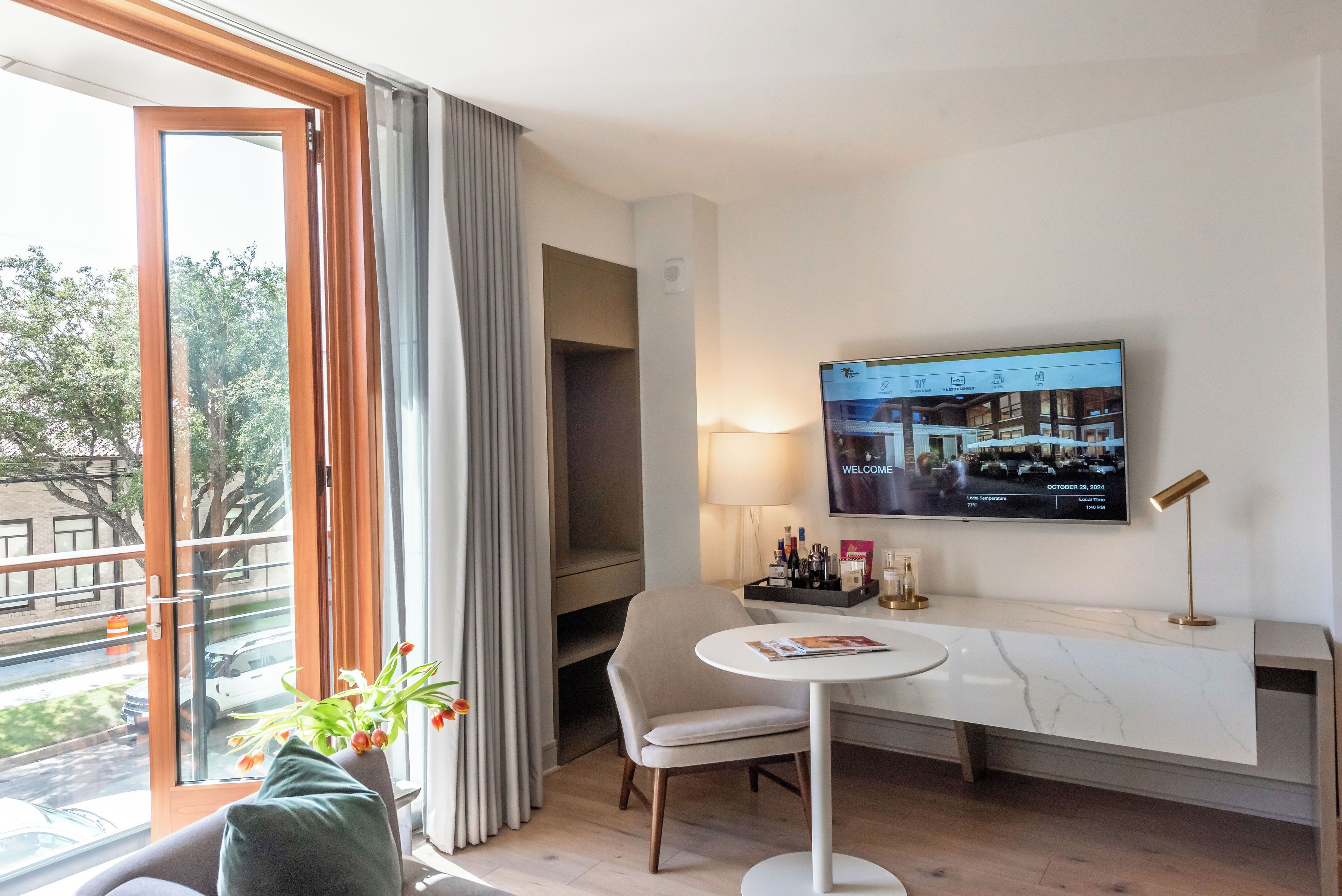 A modern hotel room with a large window, a flat-screen TV mounted on the wall, a round table with a magazine, and a comfortable chair.