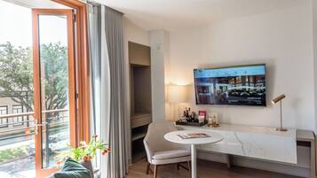 A modern hotel room with a large window, a flat-screen TV mounted on the wall, a round table with a magazine, and a comfortable chair.