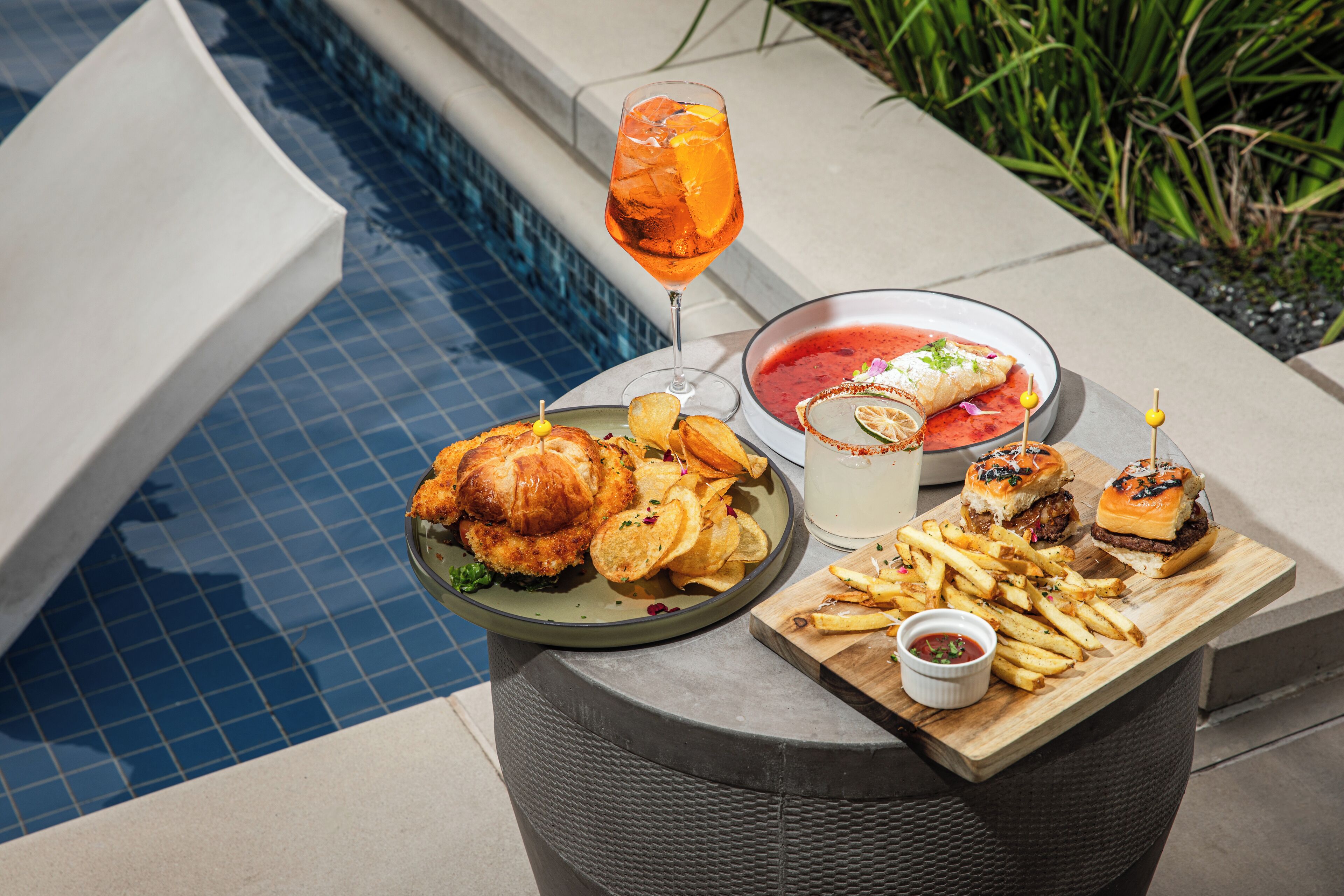 Et spisebord med et udvalg af mad, herunder en burger, pommes frites og et glas drik ved en pool.