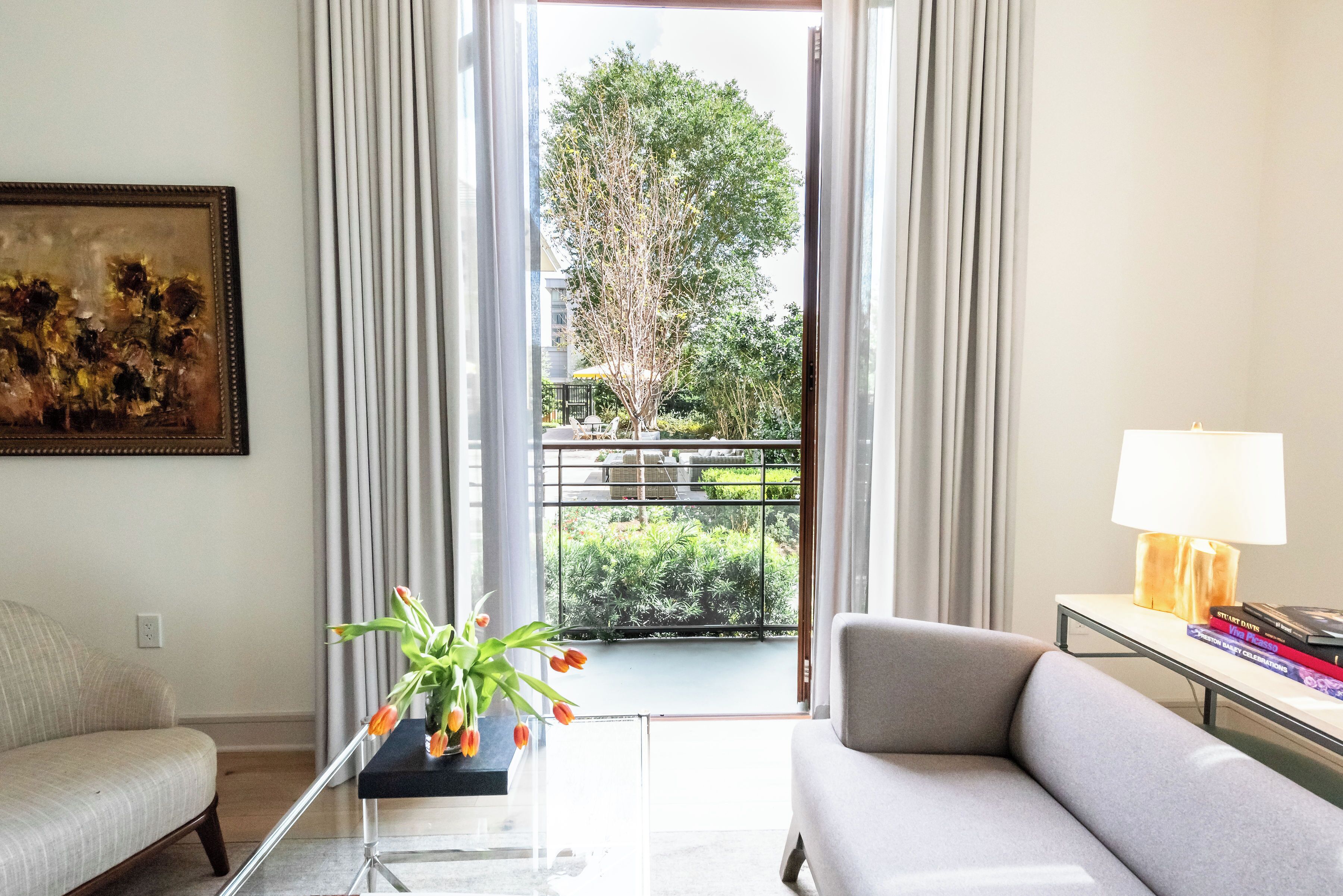 Een woonkamer met een bank, een salontafel, een schilderij aan de muur, een raam met gordijnen en uitzicht op een balkon met groen.