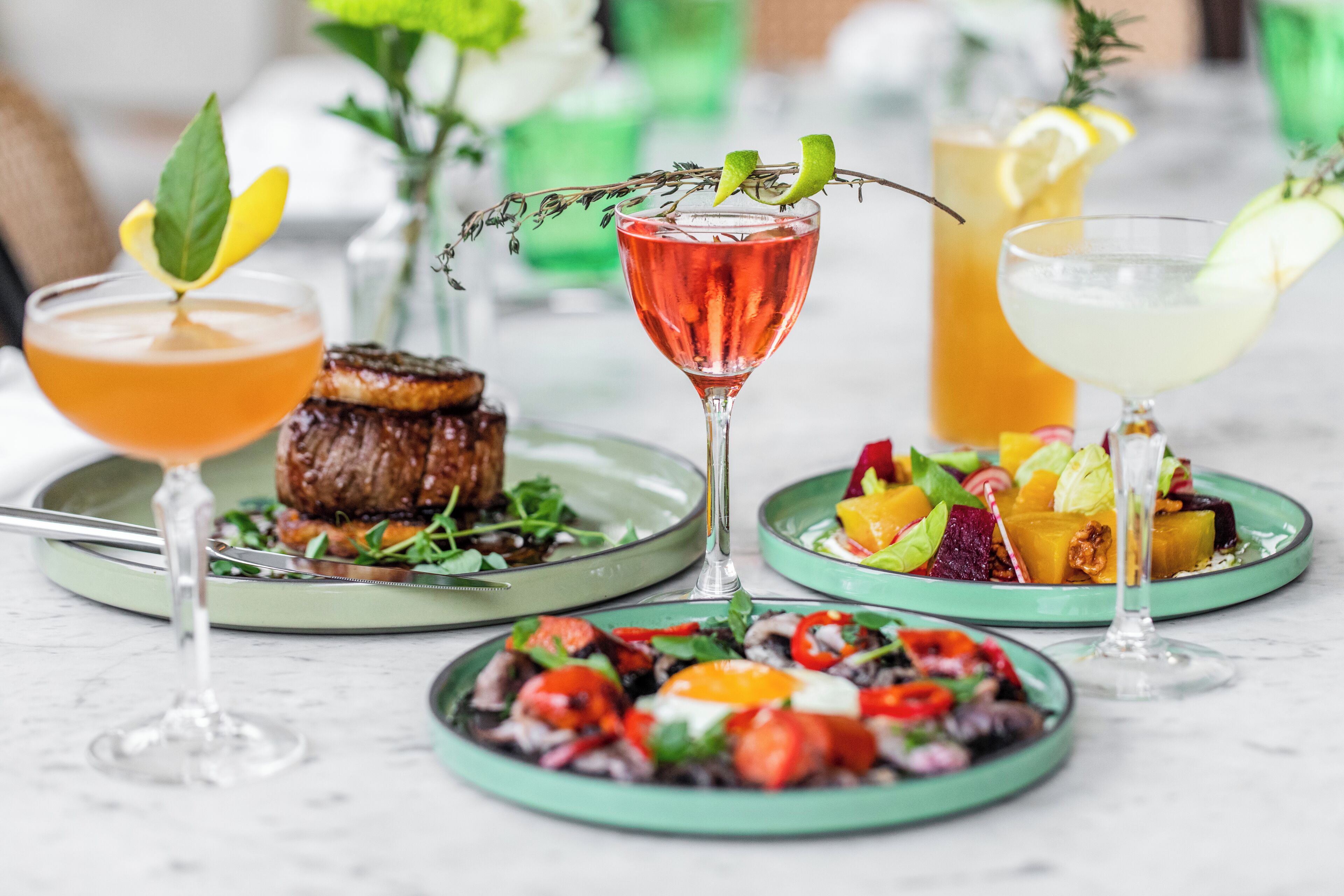 Una mesa de comedor con diversos platos y bebidas, como ensalada, filete y cócteles.