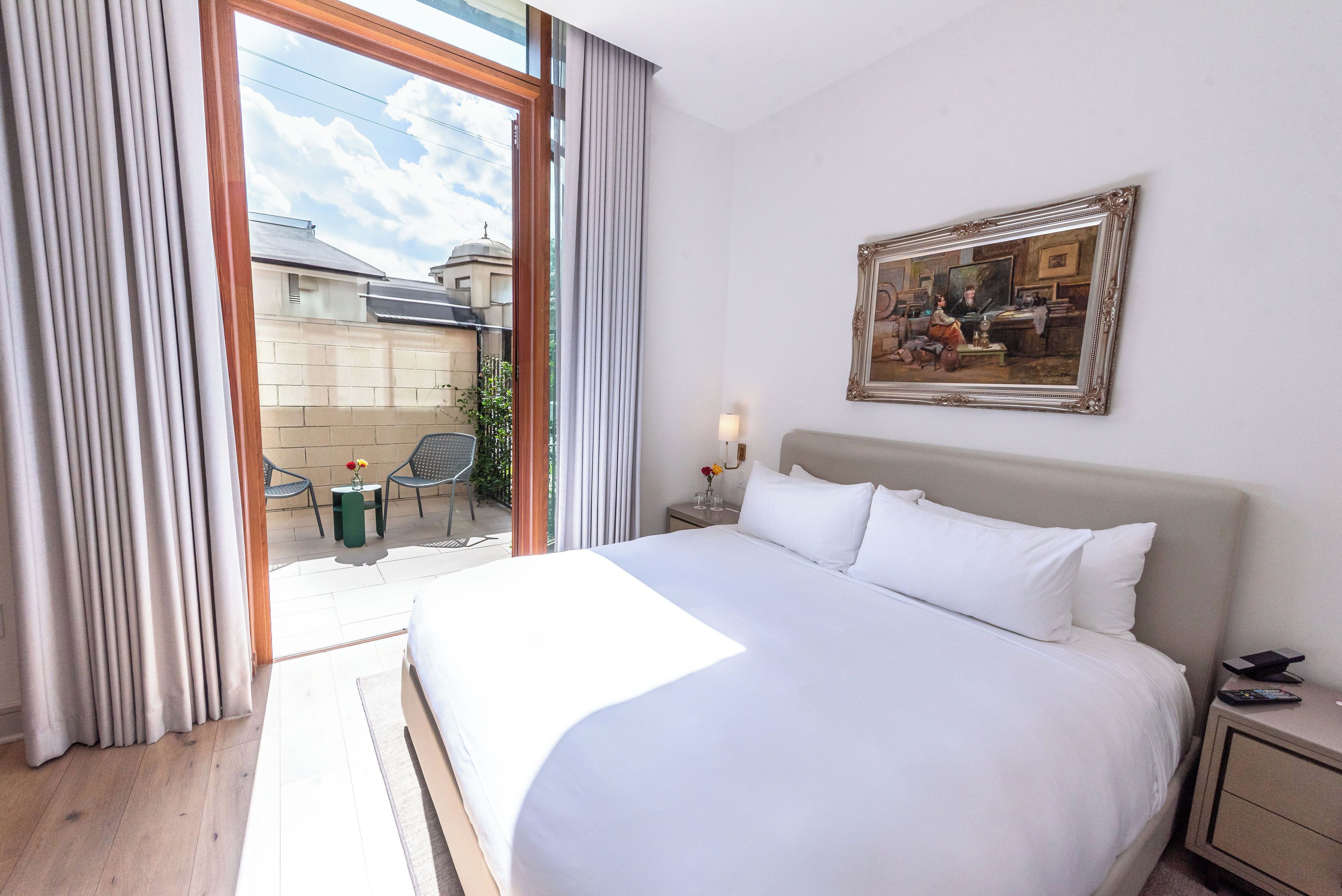 A hotel room with a large bed, a nightstand, a bedside table with a lamp, a framed picture on the wall, and a view of the outside balcony.